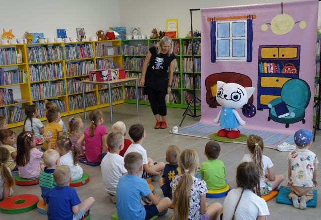 dzieci siedzą na pufach, przed nimi kobieta i maskotka kici koci