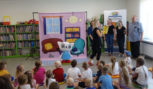 animatorka i 4 dorosłe osoby stoją przed siedzącymi na podłodze dziećmi, w tle maskorka Kici Koci i plansza z jej pokojem 