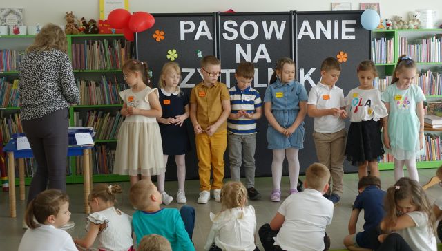 7 przedszkolaków stoi przed innymi siedzącymi na dywanie