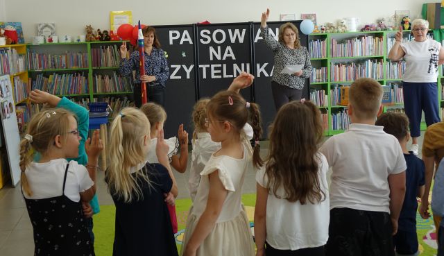 kobieta z dużą kredką i kobieta z podniesioną do góry prawą ręką stoją przed stojącymi przedszkolakami