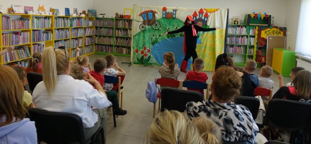 publiczność złożona z dzieci i rodziców ogląda spektakl. Kot w butach stoi przed nimi z rozłożonymi rękami, w tle plansza z jeziorem i regały z książkami