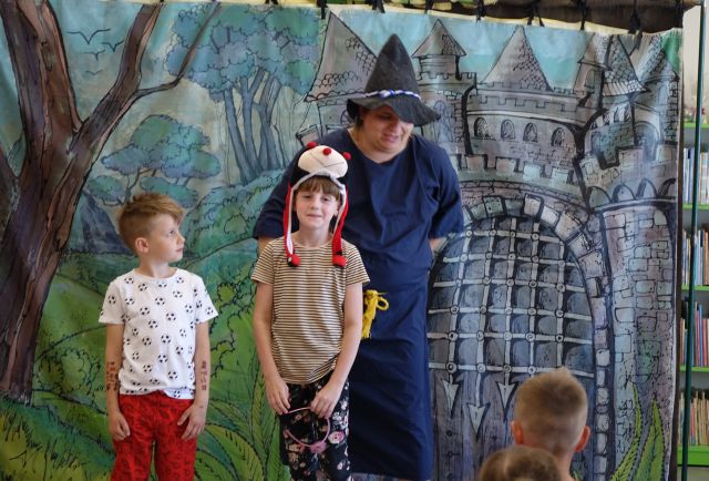 aktor przebrany za magika stoi za chłopcem i dziewczynką w czapce przedstawiającą biedronkę, w tle plansza z zamkiem