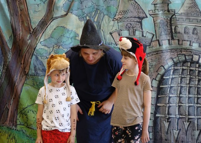 aktor przebrany za magika nachyla się nad dziećmi - chłopcem w czapce lwa i dziewczynkom w czapce biedronki, w tle zamek