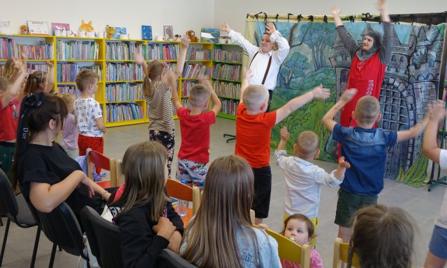aktorzy kot w butach i młynarz ćwiczą z dziećmi z podniesionymi rękoma, w tle regały z książkami