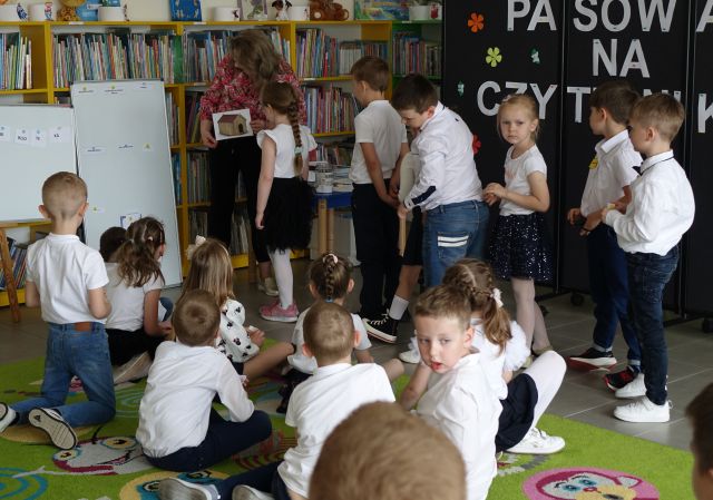6 dzieci stoi na środku sali i rozwiązuje zagadki, obok nich stoi bibliotekarka, na kolorowym dywanie siedzą pozostałe dzieci