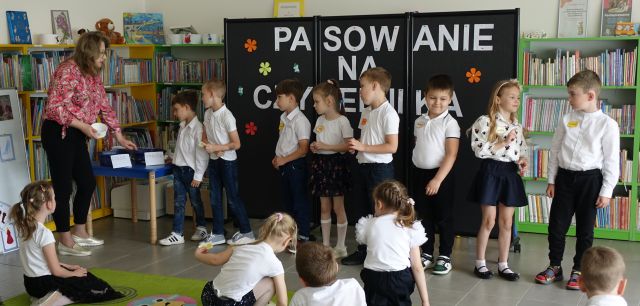 8 dzieci stoi na środku sali i rozwiązuje zagadki, przed nimi stoi bibliotekarka, pozostałe dzieci siedzą na kolorowym dywanie