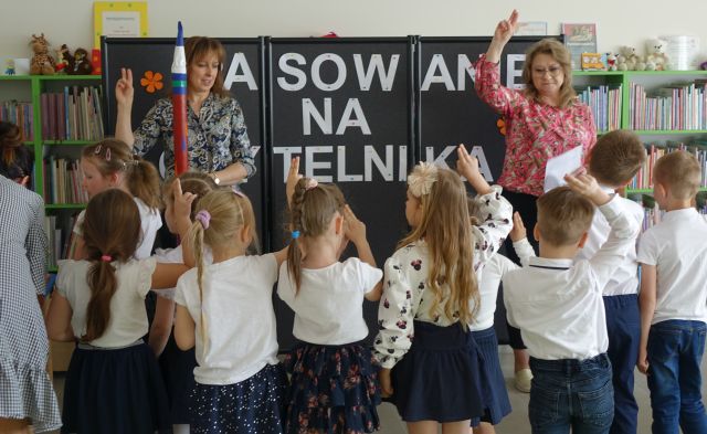 dzieci składają przysięgę stojąc, przed nimi stoi kobieta z dużą kredką i kobieta z podniesioną ręjką do góry