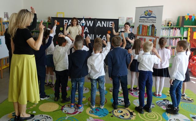 przedszkolaki i 3 kobiety stoją z podniesionymi do góry prawymi rękoma