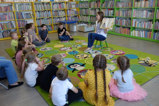 dzieci i rodzice siedzą w okręgu na kolorowym dywanie, przed nimi na krzesełku siedzi kobieta z książką, w tle regały z książkami
