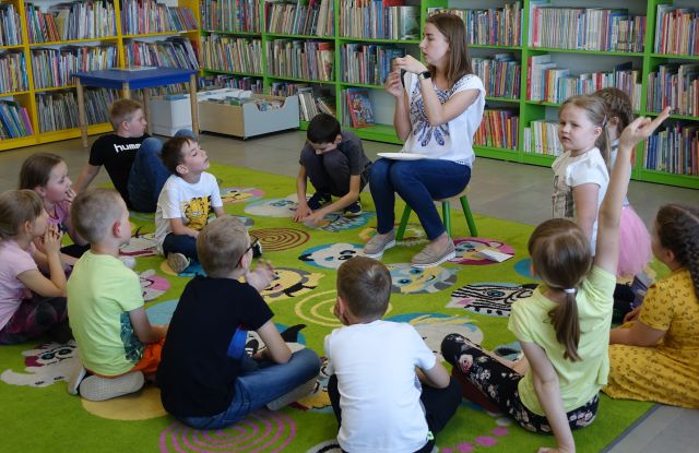 kobieta siedząca na krzesełku pokazuje dzieciom siedzącym na kolorowym dywanie rysunki owoców i warzyw