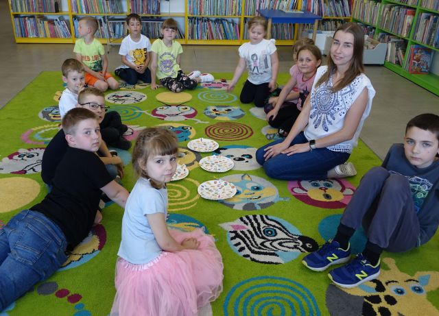 do zdjęcia pozują dzieci i kobieta siedzące na kolorowym dywanie, w tle regały z książkami