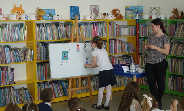 dziewczynka przypina obrazek do tablicy magnetycznej, obok stoi bibliotekarka