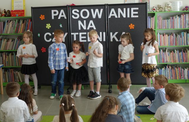 6 dzieci stoi w rzędzie przed siedzącymi na dywanie dziećmi