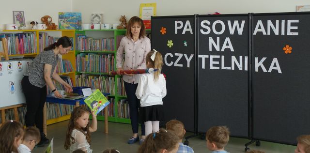dyrektorka biblioteki pasują ogromną tekturową kredką dziewczynkę na czytelniczję, obok stoi pochylona nad stolikiem bibliotekarka