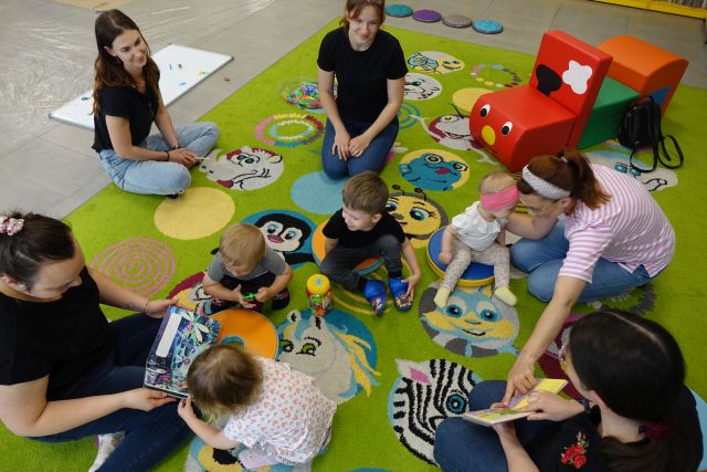 malutkie dzieci z mamusiami siedzą na kolorowym dywanie i oglądają kolorowe książeczki
