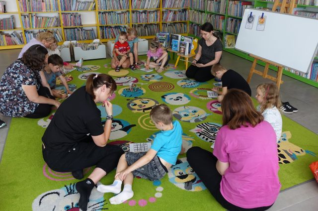 dzieci z mamami siedzą na kolorowym dywanie i wypełniają bingo, w tle regały z książkami