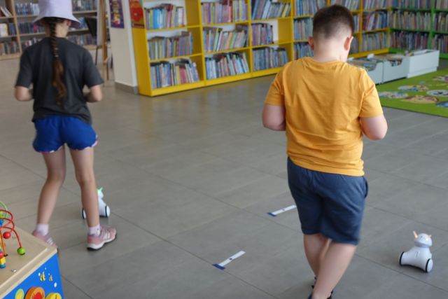 chłopiec i dziewczynka sterują robotami Photon przy użyciu tabletów, w tle regały z książkami