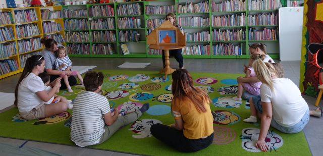 małę dzieci i rodzice siedzą na kolorowym dywanie, przed nimi na krzesełku siedzi bibliotekarka i pokazuje teatrzyk kamishibai, w tle regały z książkami
