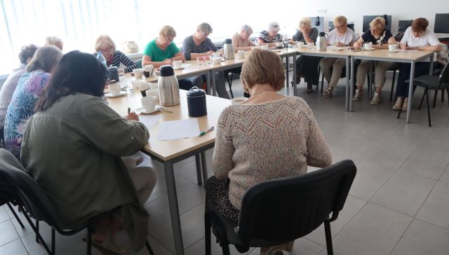 14 seniorek siedzi przy stołach ustawionych w podkowę i notuje na kartkach