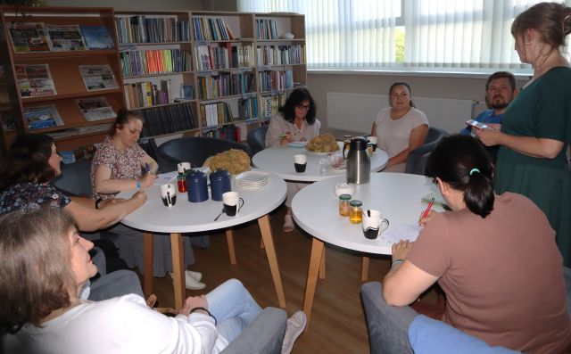 uczestnicy spotkania siedzą przy białych okrągłych stołach, w tle regały z książkami