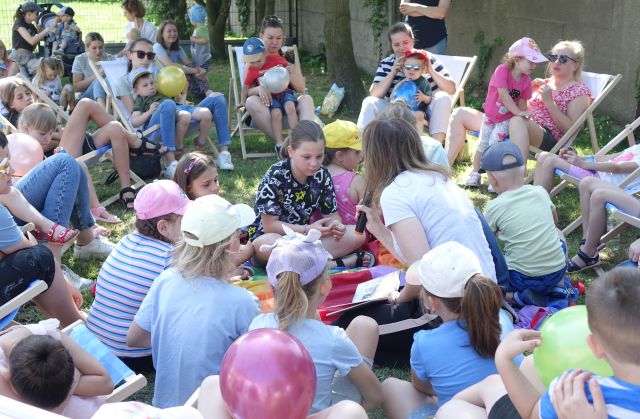dzieci i rodzice siedzący na leżakach i kolorowej chuście animacyjnej słuchają czytającej książkę kobiety