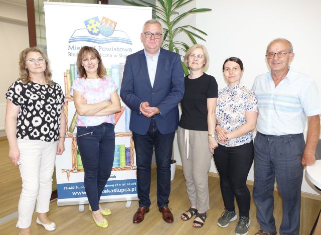 załoga biblioteki: 4 bibliotekarki i mężczyzna pozują do zdjęcia z satyrykiem, w tle baner z logo i nazwą biblioteki