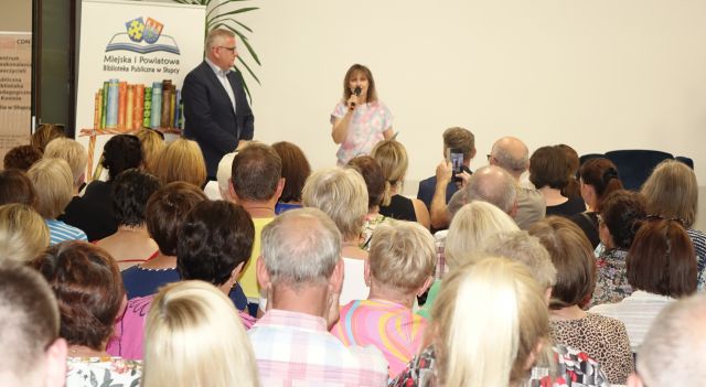 dyrektorka biblioteki z mikrofonem i mężczyzna w marynarce stoją przed liczną publicznością siedzącą na krzesłach