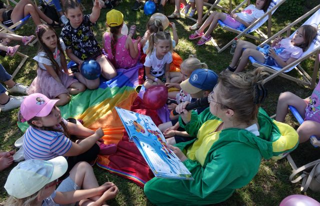 dzieci i rodzice siedzący na trawie słuchają kobiety przebranej za żabę czytającej książkę