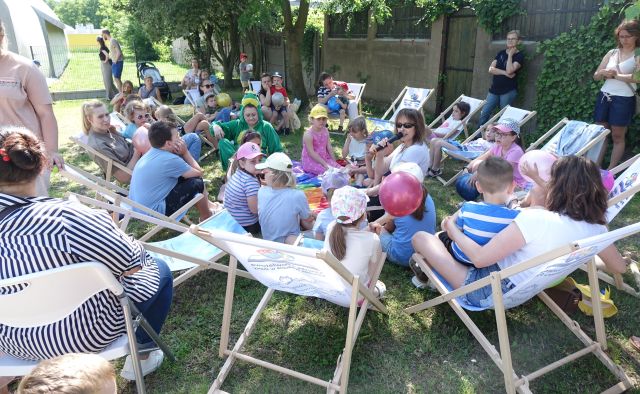 kobieta z mikrofonem i dzieci i rodzice siedzący na leżakach i kolorowej chuście animacyjnej rozłożonej na trawie