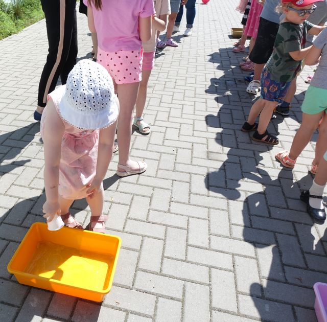dziewczynka wlewa plastikowym kubeczkiem wodę do miski, za nią w kolejce inne dzieci