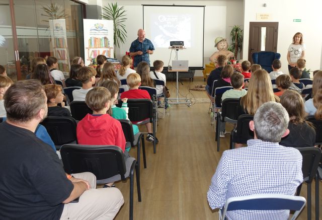 dzieci uczestniczące w spotkaniu, przed nimi stojący autor i dyrektorka biblioteki, w tle ekran i baner z logo i nazwą biblioteki