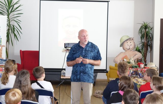 mężczyzna opowiada siedzącym przed nim na krzesłach dzieciom, w tle wyświetlana prezentacja na ekranie
