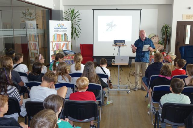 mężczyzna stojący przed siedzącymi dziećmi czyta z kartek, w tle ekran i baner z logo biblioteki