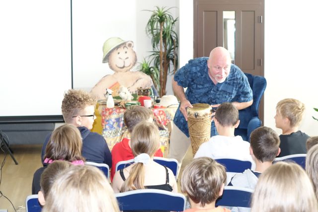 mężczyzna z bębnem pochyla się nad siedzącymi dziećmi, w tle figura niedżwiedzia w kasku