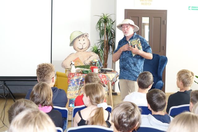 mężczyzna w czapce i z książką pod pachą stoi przed siedzącymi dziećmi