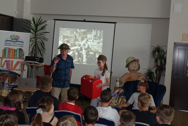 przed siedzącymi na krzesłach dziećmi stoi mężczyzna w kasku, obok stoi dziewczynka w stroju rekina oraz siedzi na fotelu chłopiec w czapce prezentującej niedżwiedzia