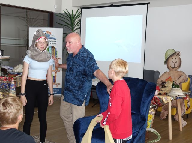 z prawej strony mężczyzny stoi chłopiec w czerwonej bluzce, po lewej dziewczynka z maską rekina