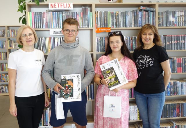 na zdjęciu z prawej strony dyrektorka biblioteki, z lewej bibliotekarka, w środku chłopak i dziewczyna z nagrodami, w tle regały z książkami