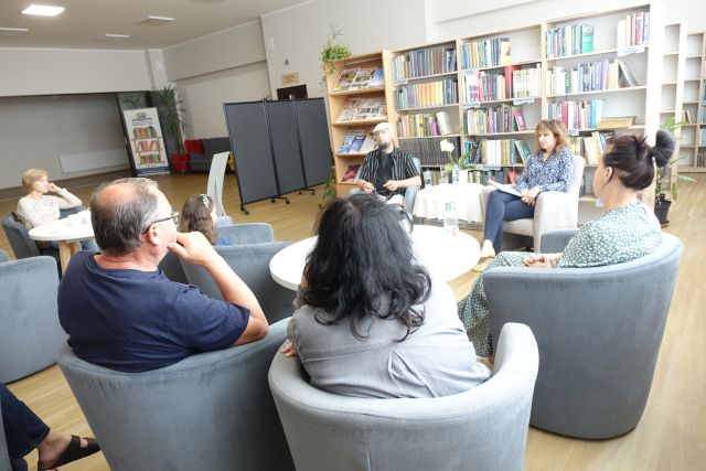 uczestnicy spotkania siedzą na fotelach przy okrągłych stołach, przed nimi muzyk i dyrektorka biblioteki, w tle regały z książkami