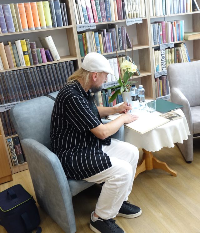 muzyk przy stoliku wpisuje się do kroniki biblioteki, w tle regały z książkami