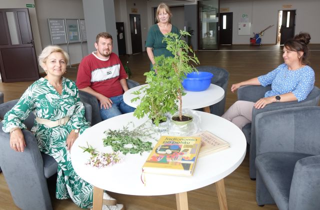 dwie kobiety i mężczyzna siedzą przy białych okrągłych stołach, jedna kobieta stoi, na jednym ze stołów roślina w doniczce oraz dwei książki