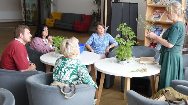 3 kobiety i mężczyzna siedzą przy białych okrągłych stołach, jedna kobieta stoi, w tle regały z książkami