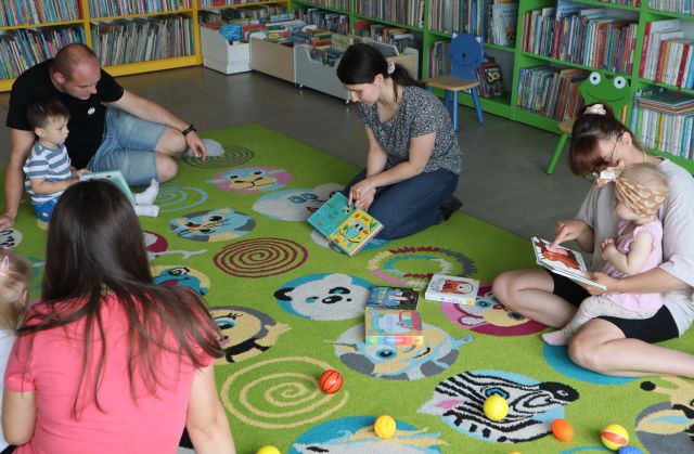 bibliotekarka pokazuje małym dzieciom i rodzicom siedzącym na kolorowym dywanie, książki, W tle regały z książkami