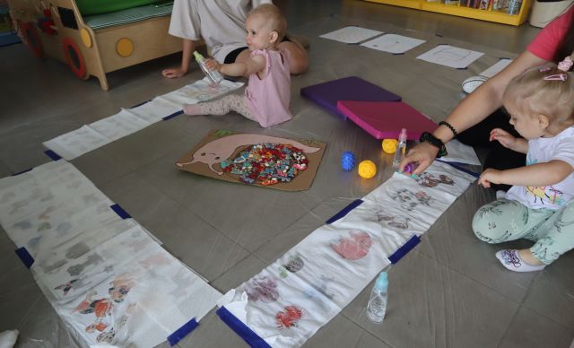 dzieci z rodzicami malują farbami na kartkach ułożonych w prostokąt
