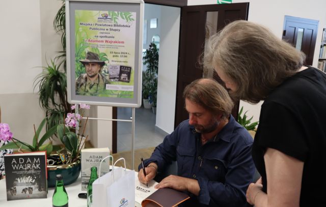 autor podpisuje książkę kobiecie, w tle plakat promujący spotkanie