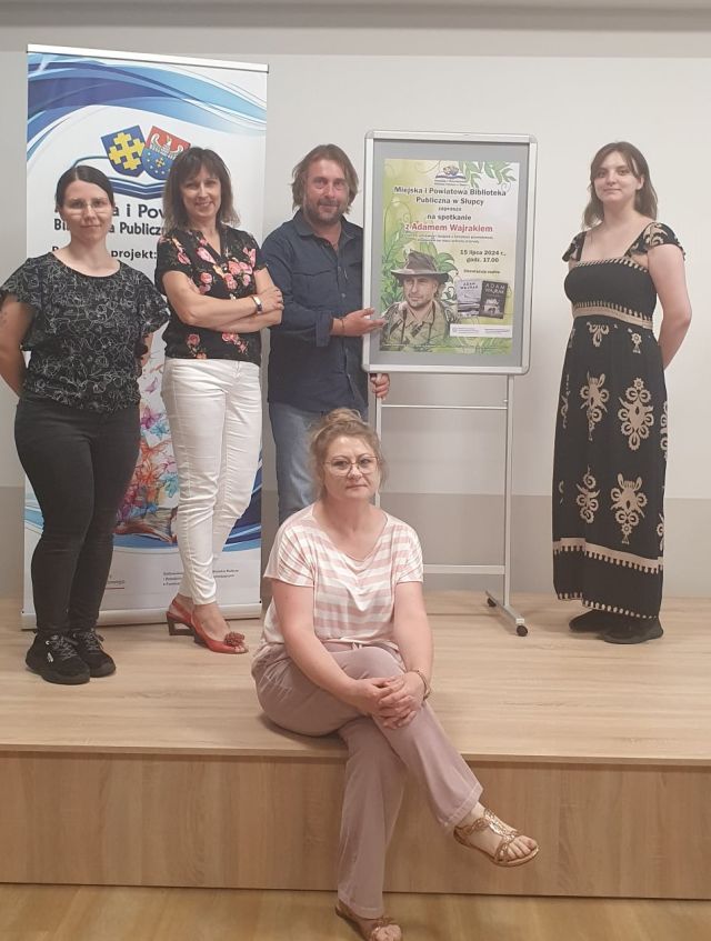 ekipa bibliotekarska pozuje do zdjęcia z autorem, z prawej strony plakat promujący spotkanie, z lewej plakat z nazwą projektu, logo biblioteki i MKiDN