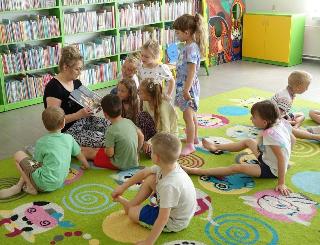 bibliotekarka siedząca na kolorowym dywanie pokazuje przedszkolakom zgromadzonym wokół niej ilustracje w książce, w tle regały z książkami