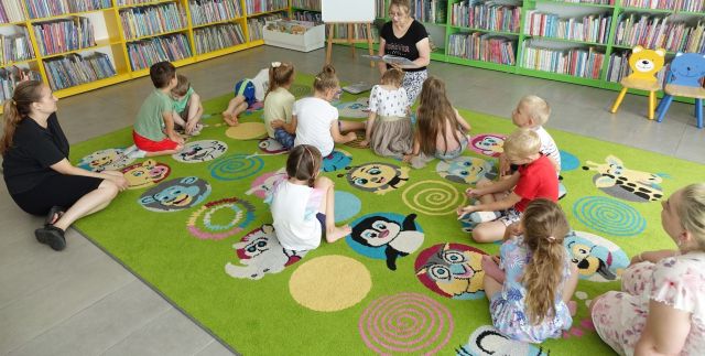 przedszkolaki siedzą na kolorowym dywanie, przed nimi siedzi bibliotekarka z książką, w tle regały z książkami