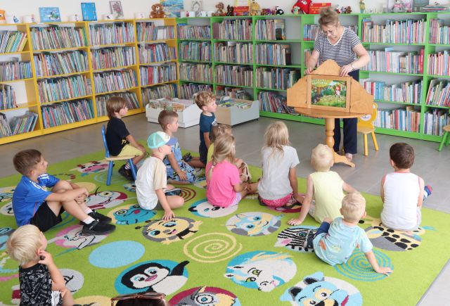 przedszkolaki siedzą na kolorowym dywanie i oglądają teatrzyk kamishibai, w tle regały z książkami