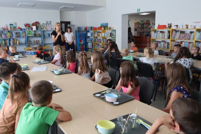 dzieci uczestniczące w warsztatach siedzą przy stolikach, słuchając opowiadającej kobiety, obok stoi dyrektorka biblioteki. W tle regały z książkami